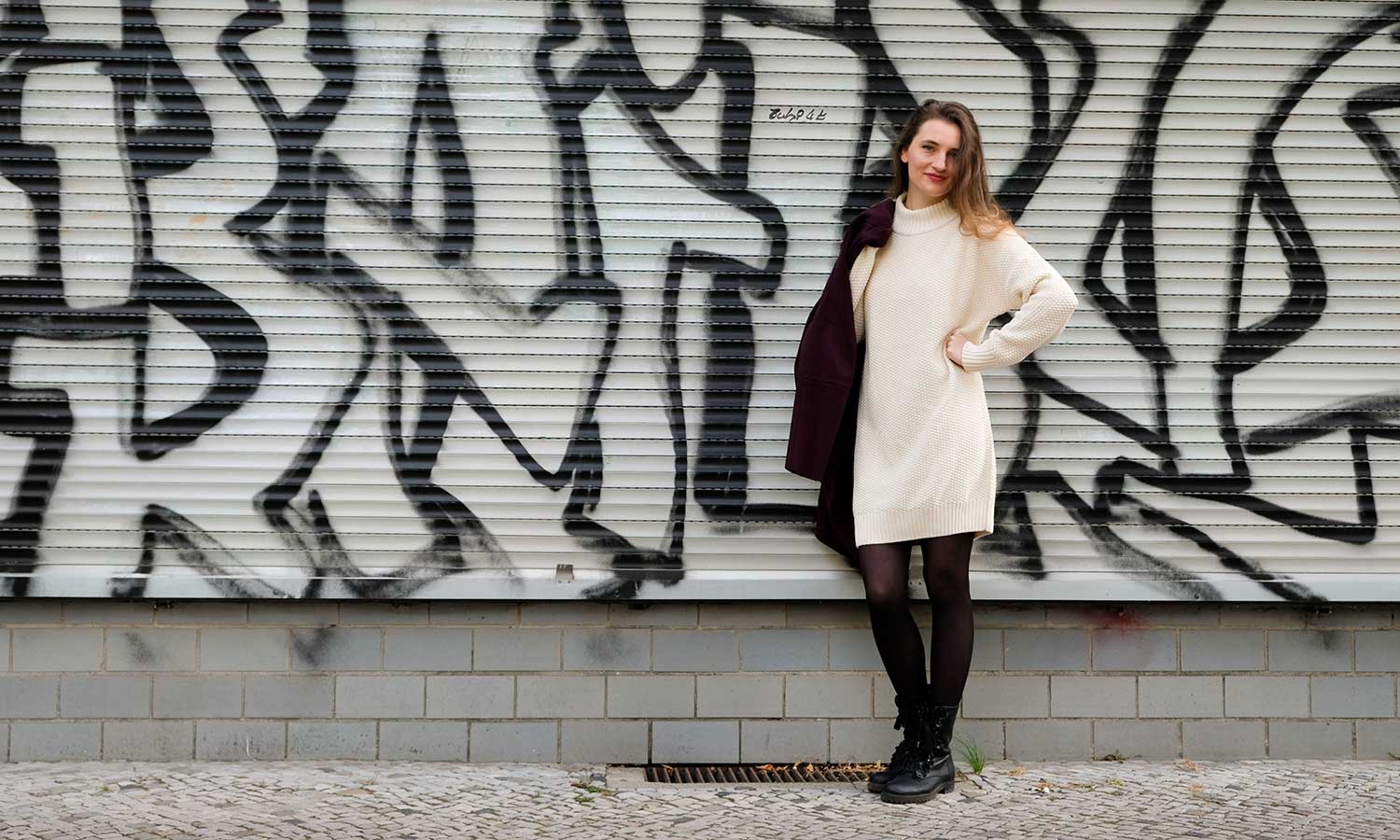 Frau in weißem Strickkleid steht vor einer Graffiti Wand