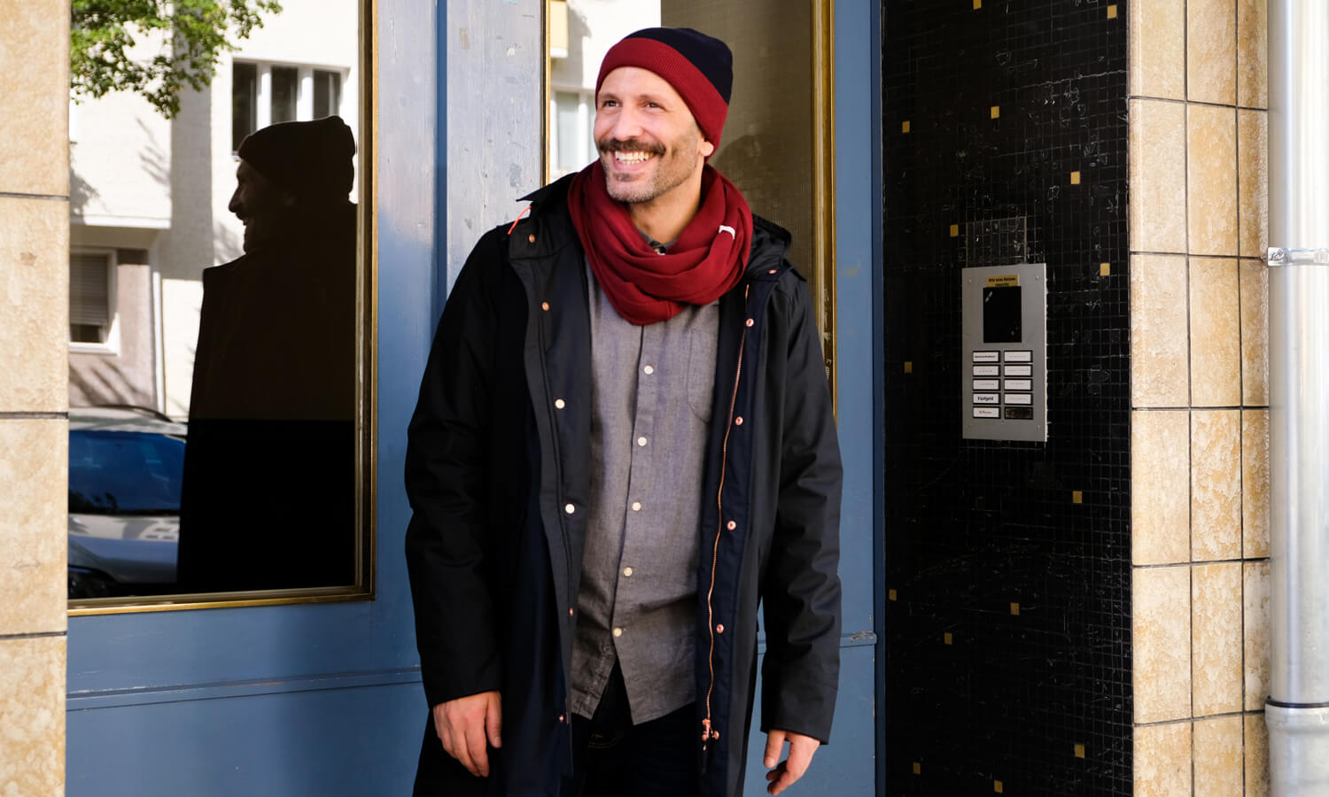Mann steht im Hauseingang und trägt schwarze Winterjacke und Strickaccessoires